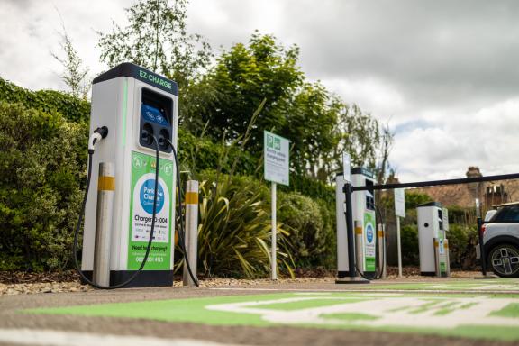 Electric vehicle charging point in a small outdoor car park, with multiple “Park & Charge Oxfordshire” chargers lined up beside greenery. A cable is plugged into the foreground charger, with EV parking markings on the ground and signage indicating an electric vehicle charging point.